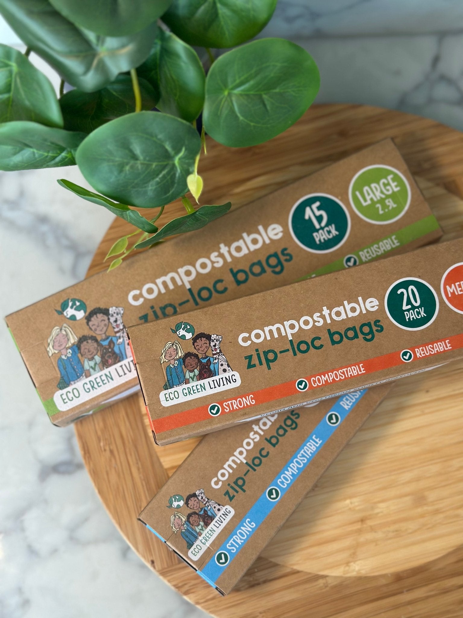 Three boxes of compostable zip lock bags in large, medium and small sizes on a wooden tray beside a green plant